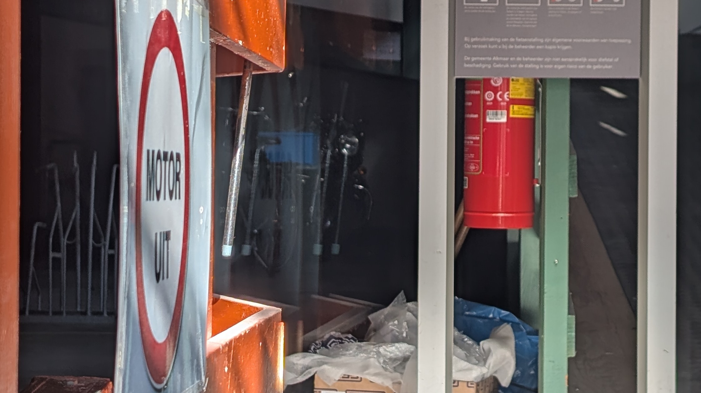 Een bord met de tekst 'MOTOR UIT' hangt naast een brandblusser in een ruimte gevuld met diverse voorwerpen en een instructieposter aan de muur.