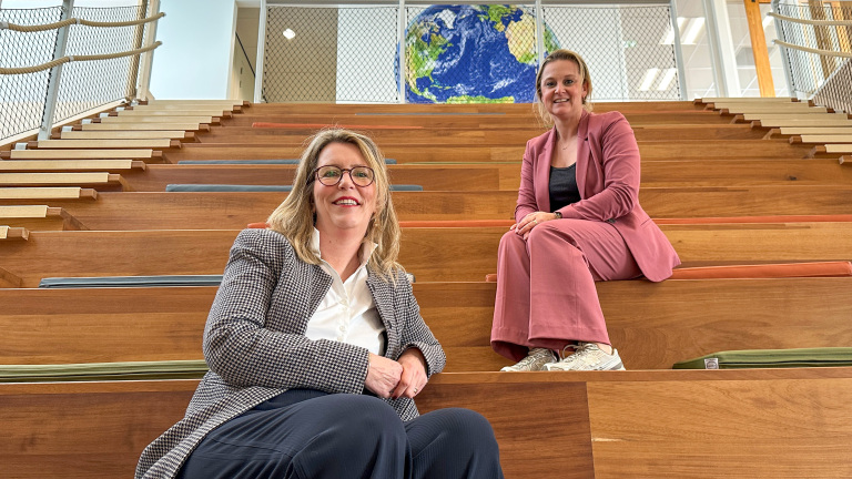 Twee vrouwen zitten op een houten trap met kleurrijke kussens in een moderne binnenruimte; op de achtergrond is een afbeelding van de aarde zichtbaar.