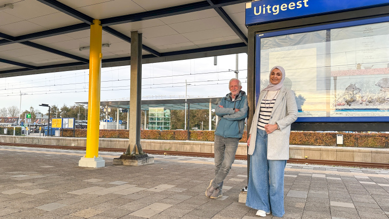 Twee mensen poseren naast een informatiebord op het treinstation Uitgeest. Eén persoon leunt tegen een gele pilaar, de ander staat ernaast. Het perron is leeg, en er zijn sporen en bushaltes te zien op de achtergrond.