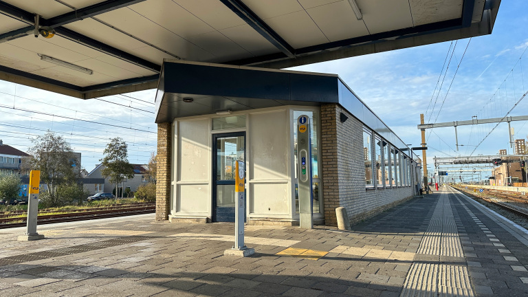 Een treinperron met een modern stationsgebouw, voorzien van witte en donkerblauwe elementen, en een toegangspoortje. Het perron is leeg en er zijn sporen zichtbaar aan de rechterkant. Het gebouw heeft een glazen deur en ramen, en er zijn informatie- en OV-kaartlezers aan de buitenkant.