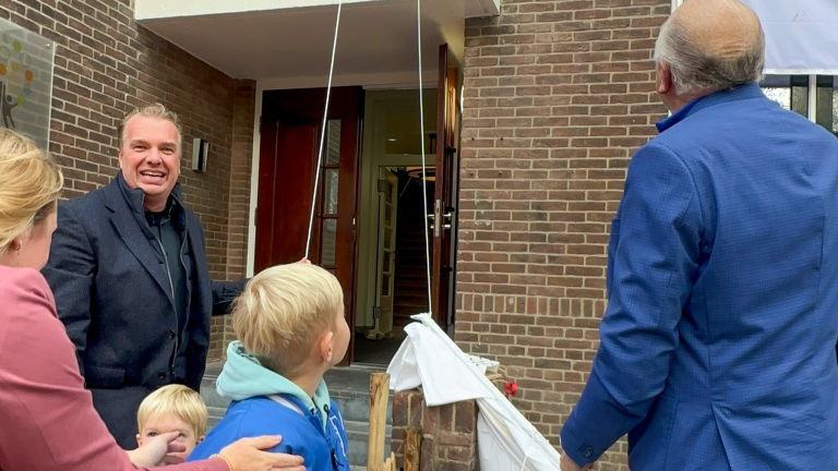 Groep mensen die samen voor een stenen gebouw staan, terwijl een kind en een volwassene een koord vasthouden.