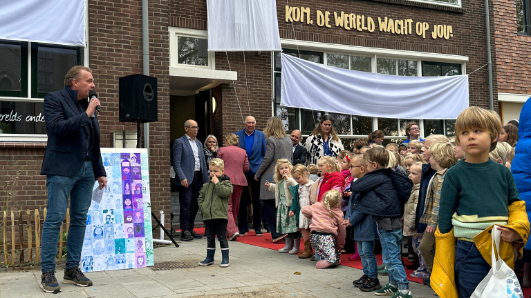 Een man spreekt in een microfoon buiten voor een gebouw met een groep kinderen en volwassenen. Op de achtergrond hangt een doek met de tekst 'Kom, de wereld wacht op jou!' op de muur. Kinderen staan op een rij voor het gebouw, sommige kijken naar de camera terwijl anderen naar de man luisteren.