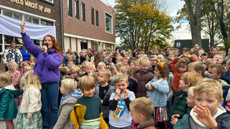 Een vrouw in een paarse jas spreekt met een microfoon voor een grote groep kinderen die buiten voor een bakstenen gebouw staan met de tekst "De wereld wacht op jou!"