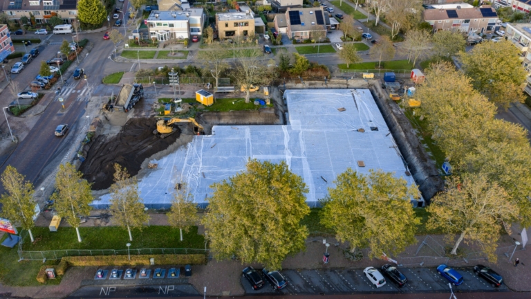 Luchtfoto van een bouwplaats in een stedelijke buurt, waarbij een groot deel is bedekt met witte zeilen. Een graafmachine is bezig met het verplaatsen van zand aan de linkerkant van de locatie. Er zijn bomen aan de rand van de bouwplaats en omringende woonhuizen.