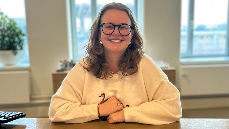 Vrouw met bril en gekruld haar lacht terwijl ze aan een houten bureau zit in een lichte kamer met grote ramen.