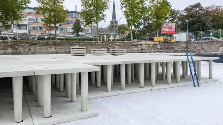 Betonnen constructie met pilaren in de bouwfase, tegen de achtergrond van een rij bomen en een kerk met een torenspits. Op de achtergrond zijn een gebouw en een hek zichtbaar. Er staat een blauw laddertje tegen de constructie aan.