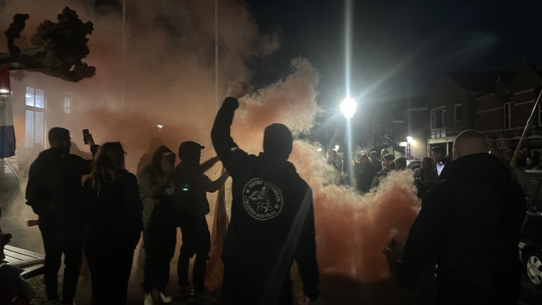 Groep mensen in de nacht bijeen in een straat, omringd door oranje rook van een fakkel, met een vlag zichtbaar en een lantaarnpaal op de achtergrond.