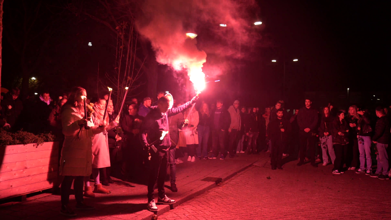 Een groep mensen staat 's nachts buiten, omringd door fakkels en een rode rookwolk, terwijl een persoon een fakkel omhoog houdt.
