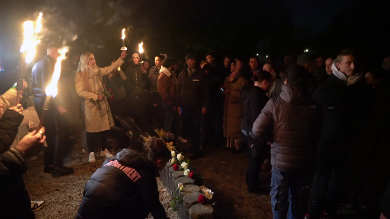 Een groep mensen verzameld in het donker, sommige met fakkels, bij een herdenkingsplek met bloemen op de grond.