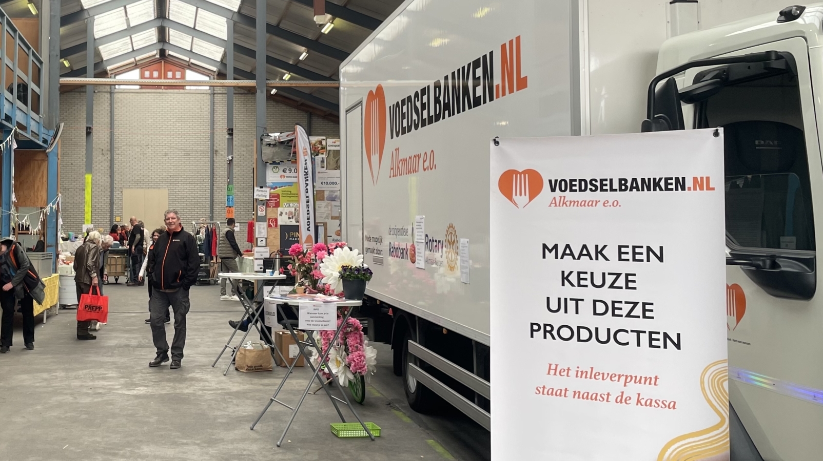 Een binnenruimte van een voedselbank in Alkmaar met een grote witte vrachtwagen en een informatiestand bij de ingang. Er staan mensen bij tafels met voedselproducten. Aan de zijkant is een houten trap zichtbaar en er hangen vlaggetjes langs de railing van het bovenste niveau. Op de voorgrond is een bord te zien met de tekst van de voedselbank.