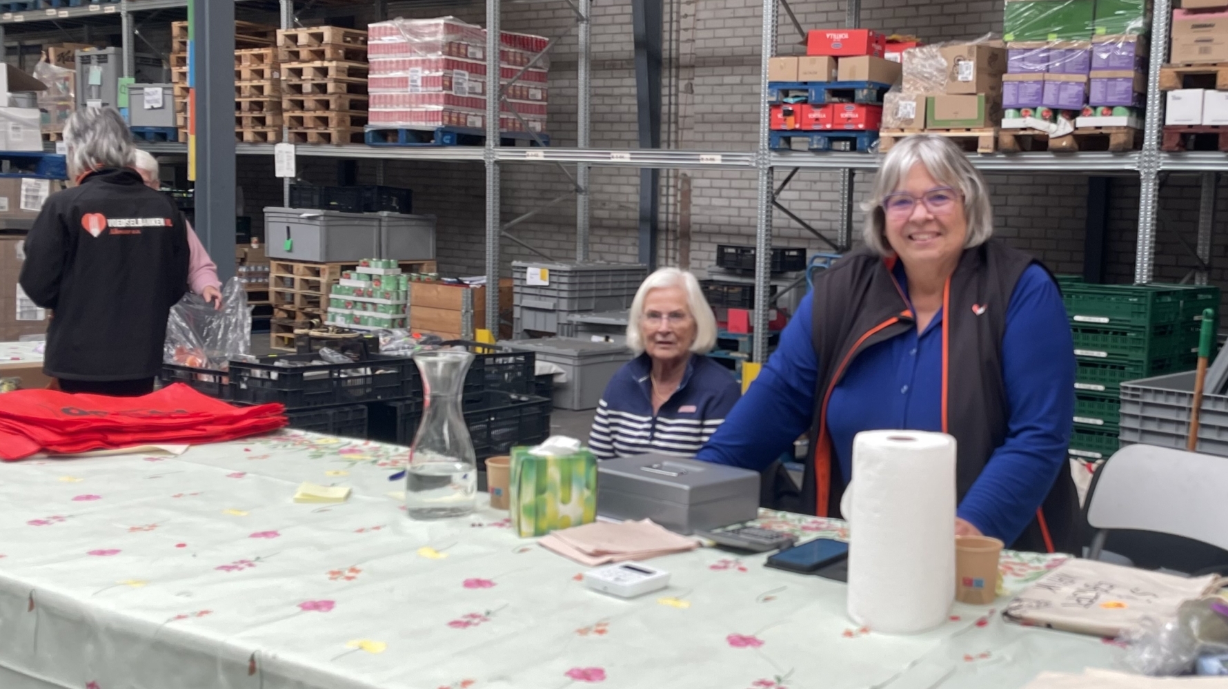 Twee vrouwen zitten aan een tafel in een magazijn, met rekken vol spullen op de achtergrond. De tafel is bedekt met een lichtgekleurd tafelkleed met bloemenpatroon. Op de tafel staan verschillende producten en een rol keukenpapier.