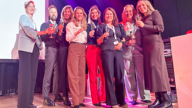 Een groep van acht lachende mensen poseert met glazen champagne in een verlichte ruimte, sommigen dragen zakelijke kleding.