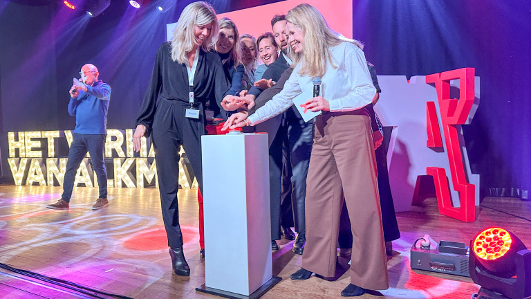 Een groep mensen staat op een podium en drukt gezamenlijk op een knop. Op de achtergrond zijn verlichte letters en een persoon met een microfoon zichtbaar. De sfeer lijkt feestelijk met gekleurde verlichting.