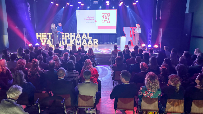 Een goed gevuld auditorium waarbij een presentatie wordt gegeven over een digitaal brandbook. Twee mensen staan op het podium naast een groot scherm, terwijl het publiek aandachtig luistert. Op de achtergrond zijn verlichte letters die "Het verhaal van Alkmaar" vormen.