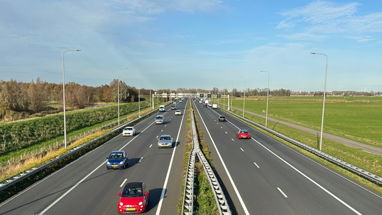 Autos rijden op een tweebaanssnelweg met groene velden en bomen aan weerszijden onder een heldere blauwe lucht.