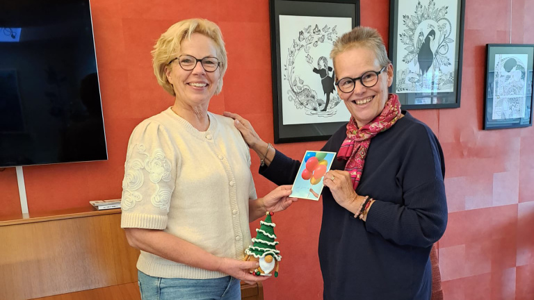 Twee vrouwen staan binnen met een kaart en een gehaakte kerstboomknuffel, glimlachend naar de camera.
