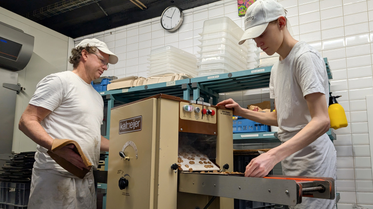 Twee bakkers in een bakkerij werken aan een machine die koekjes produceert. Eén man staat links met een beschermende handschoen en kijkt naar de machine, terwijl de jongere persoon aan de rechterkant koekjes van de transportband haalt. Op de achtergrond zijn witte tegels en een wandklok zichtbaar.