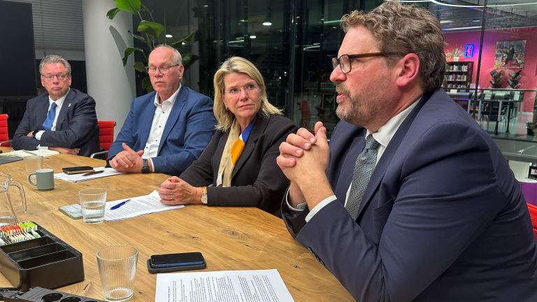 Vier mensen in zakelijke kleding zitten aan een tafel tijdens een vergadering, met notities en glazen water voor hen.