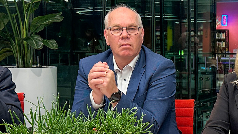 Een man met een bril en een blauw pak zit aan een tafel met gevouwen handen, omringd door planten en een moderne, verlichte achtergrond.