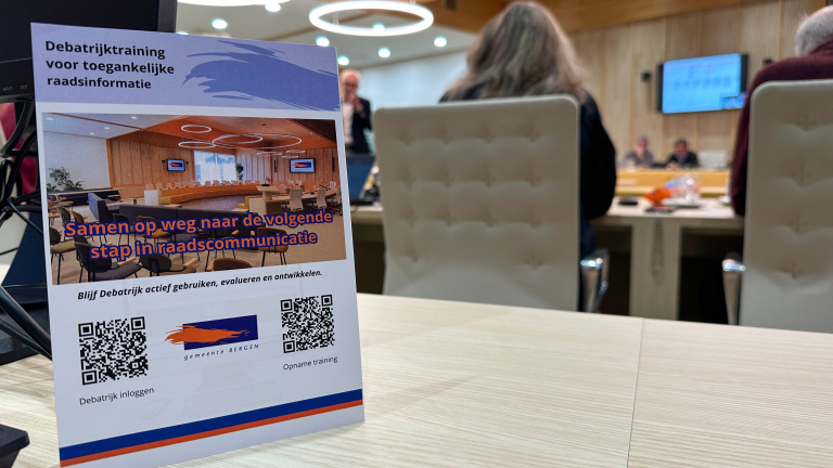 Folder met informatie over een training genaamd 'Debatrijk' voor toegankelijke raadsinformatie, met een afbeelding van een vergaderruimte en QR-codes, op een tafel in een vergaderzaal waar mensen aan een vergadering deelnemen.