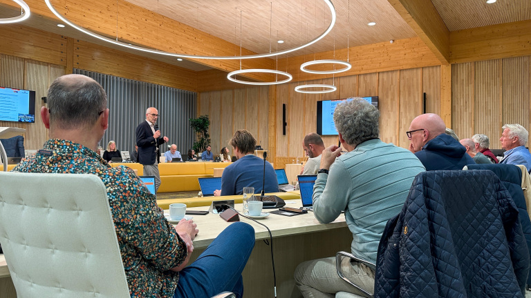 Groep mensen in een vergaderruimte met houten muren, zittend rond een tafel met laptops en papieren, terwijl een spreker met microfoon aan het presenteren is.