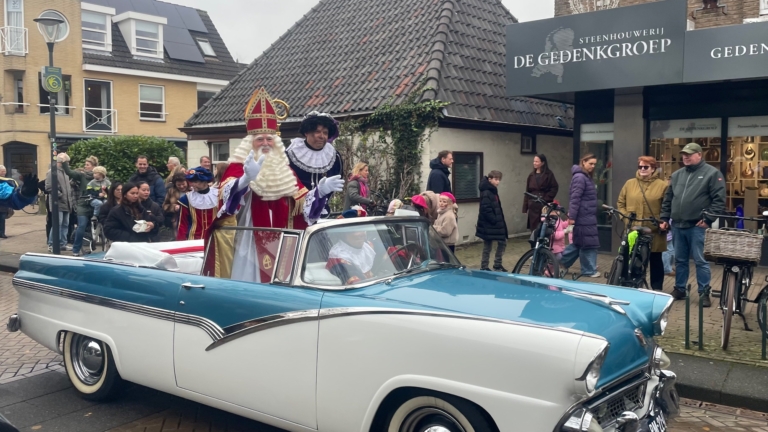 Sinterklaas en Pieten rijden in een klassieke blauwe en witte cabriolet langs een straat met toeschouwers, tijdens een Sinterklaasintocht.