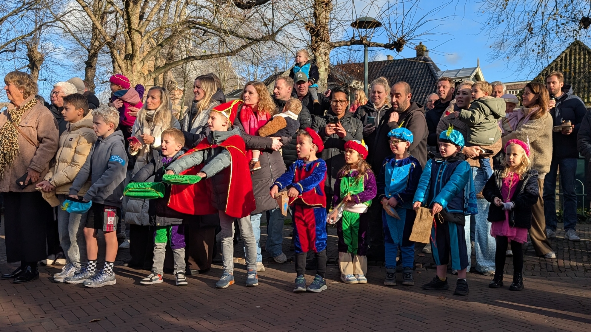 Een groep kinderen en volwassenen verzameld op een plein, sommige kinderen zijn verkleed als Zwarte Piet, terwijl anderen warme winterkleding dragen; de achtergrond toont kale bomen en een blauwe lucht.