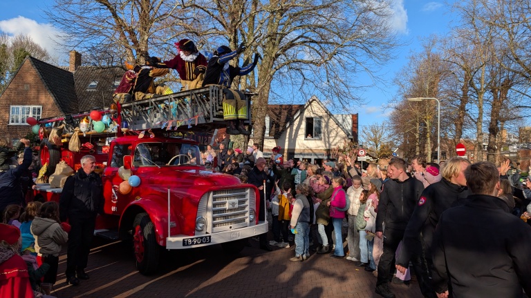 Een kleurrijke parade met een rode oude brandweerwagen waarop Pieten staan, die zwaaien naar een menigte van mensen, waaronder veel kinderen, op een zonnige dag in een bestraat dorp met bomen en huizen op de achtergrond.
