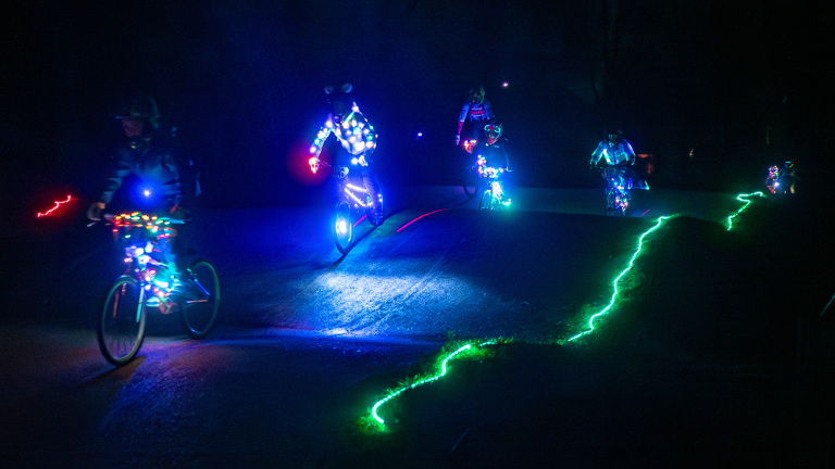 Groep fietsers rijdt 's nachts op een verlichte baan, met felgekleurde lichten op hun kleding en fietsen.