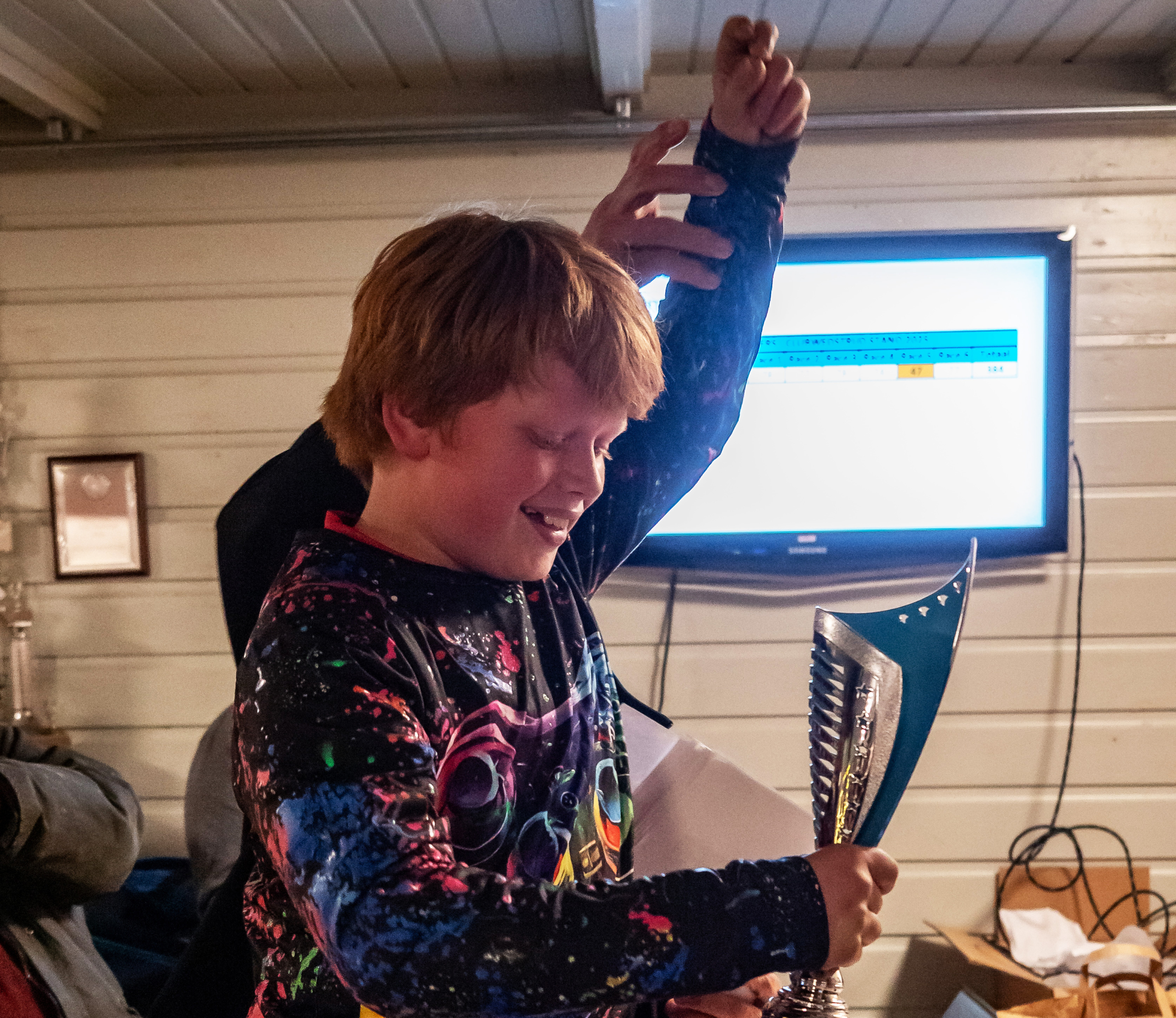 Een kind met rood haar in een kleurrijke trui houdt een trofee omhoog, terwijl hij blij kijkt in een interieur met een televisie op de achtergrond.
