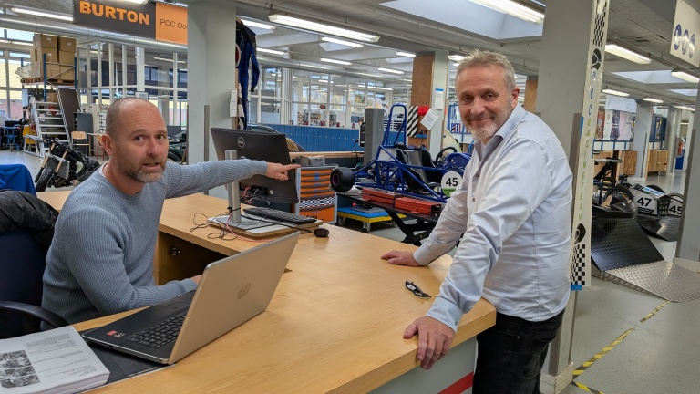 Twee mannen staan bij een balie in een werkplaats, met technische apparatuur en gedeeltelijk gemonteerde voertuigen op de achtergrond. Eén man wijst naar een computerscherm terwijl de andere glimlachend naar de camera kijkt.
