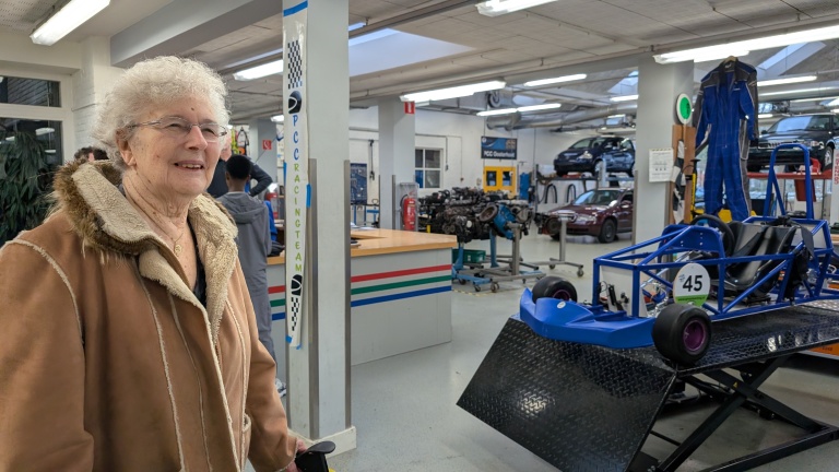 Een oudere vrouw met een bril en een bruine jas staat in een werkplaats met een kart op een platform en diverse autoonderdelen op de achtergrond.