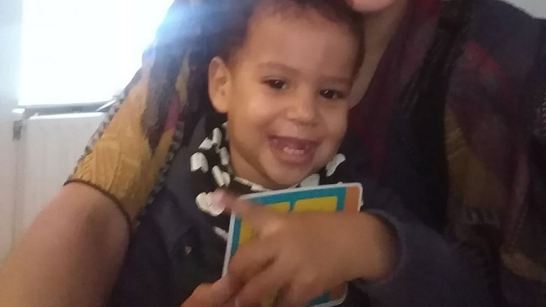 Een lachende peuter houdt een kaart vast, zittend op de schoot van een volwassene in kleurrijke kleding.