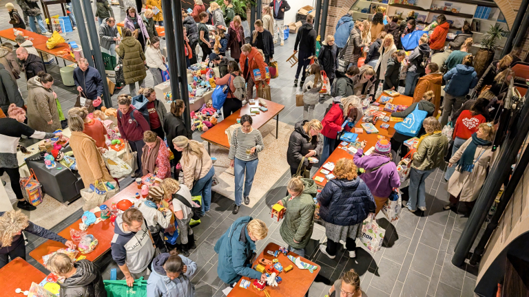 Een drukbezochte ruimte met veel mensen die rond verschillende tafels met speelgoed en huishoudelijke artikelen lopen.