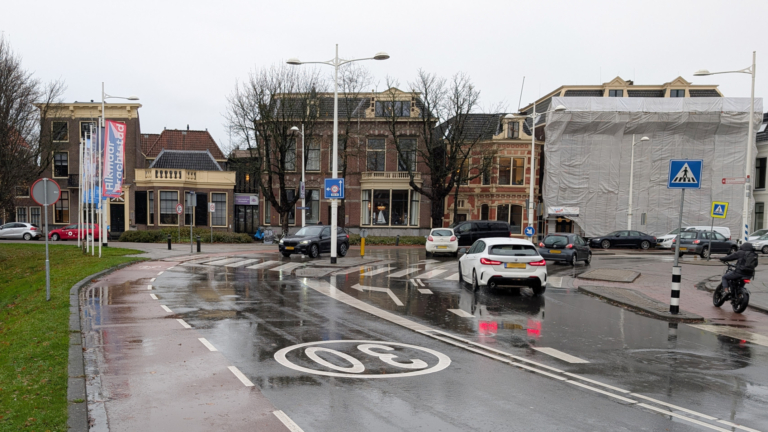 Een natte straat in een woonwijk met historische gebouwen, auto's en een fietser, met verkeersborden en een 30 km/u markering op de grond.