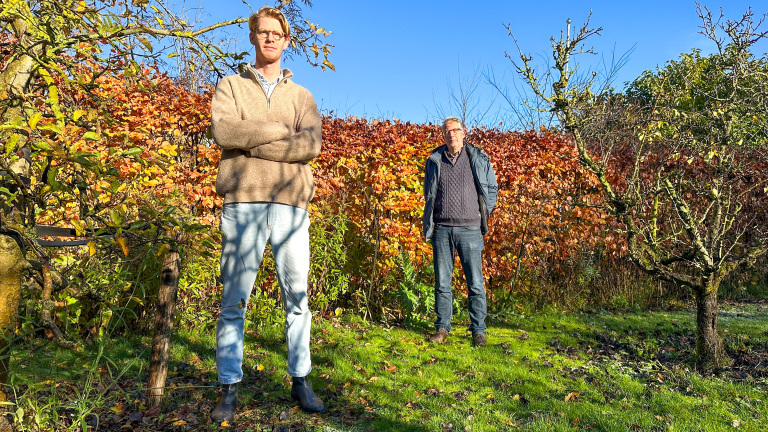 Twee mannen staan in een herfsttuin met oranje bladeren op de achtergrond, een man is op de voorgrond met gekruiste armen en de ander staat erachter. Ze dragen casual kleding en het weer is zonnig.