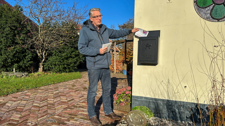 Een man in een blauwe jas en jeans steekt een brief in een zwarte brievenbus bevestigd aan een muur, met een betegeld pad en tuin op de achtergrond.