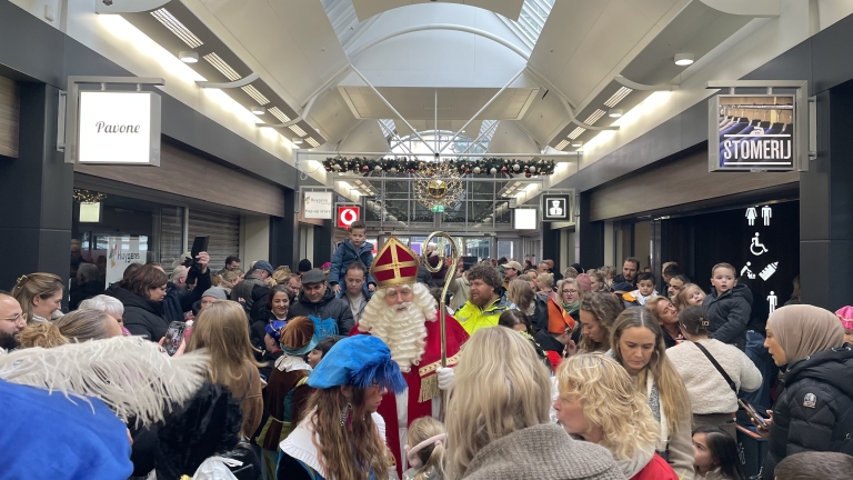 Een drukke winkelpassage met Sinterklaas in het midden, omringd door volwassenen en kinderen tijdens een evenement. Versieringen hangen aan het plafond en een kind zit op de schouders van een volwassene.