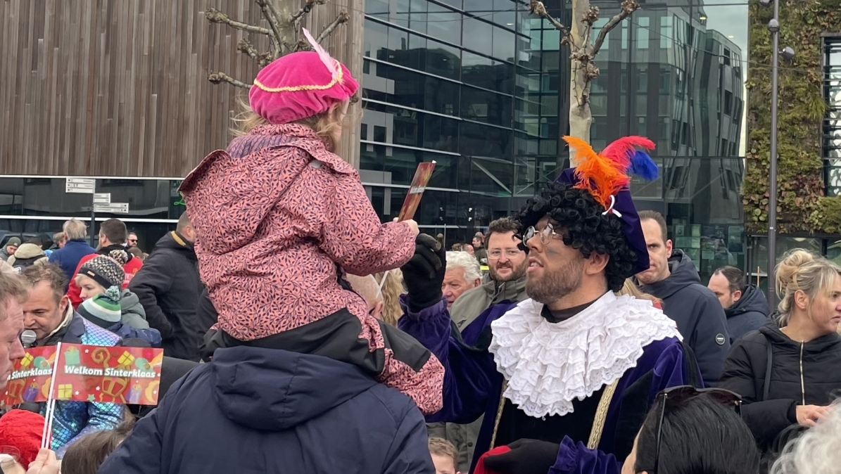 Een menigte mensen verzameld op een plein, met een persoon gekleed als een traditionele feestfigurant met een gekleurd kostuum en een kind dat op schouders zit met een roze muts op, omringd door moderne gebouwen.