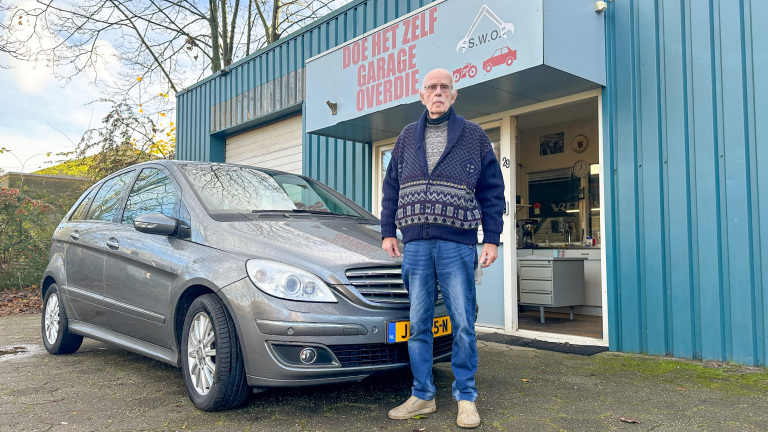 Een man staat voor een grijze auto en een garage met het bord 'Doe Het Zelf Garage Overdie'.