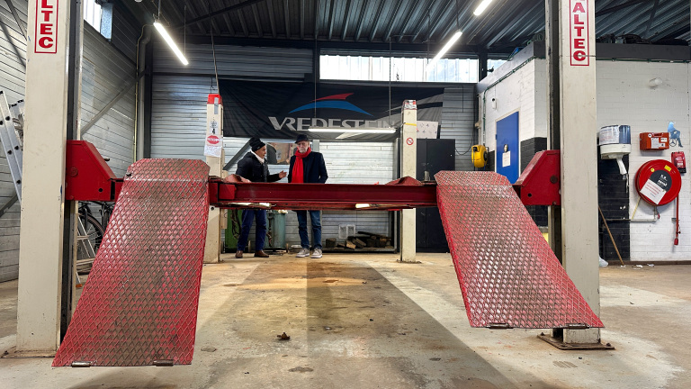 Twee mensen staan in een autowerkplaats naast een rode autolift van het merk Altec, met een Vredestein bord op de achtergrond.