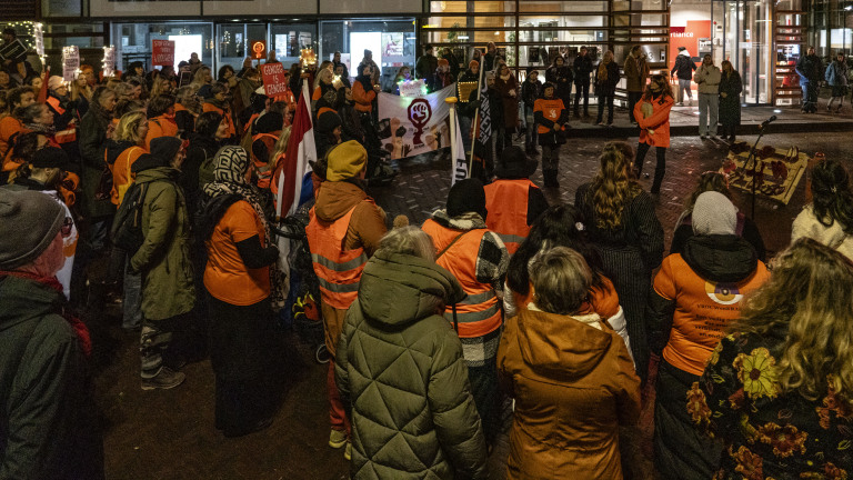 Een menigte mensen gekleed in oranje en winterjassen verzameld voor een protest tegen geweld, met spandoeken en borden in een stadsomgeving.