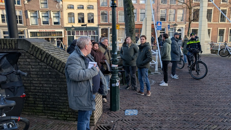 Een groep mensen staat op een met klinkers geplaveide straat in een stad, met op de achtergrond grachtenpanden en een brug. Een van de mensen is een politieagent op een fiets.