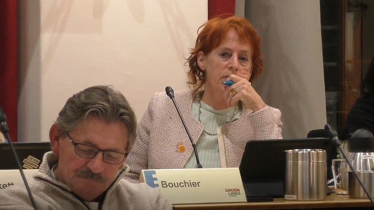 Een vrouw met roodbruin haar in een vergadering, met een microfoon voor haar, naast een man met een bril. Er staat "Bouchier" en "GroenLinks" op een naambordje voor haar.