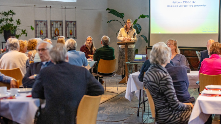 Een vrouw geeft een presentatie aan een groep mensen in een zaal, met op de achtergrond een projectiescherm waarop 'Heiloo – 1960-1965' staat.