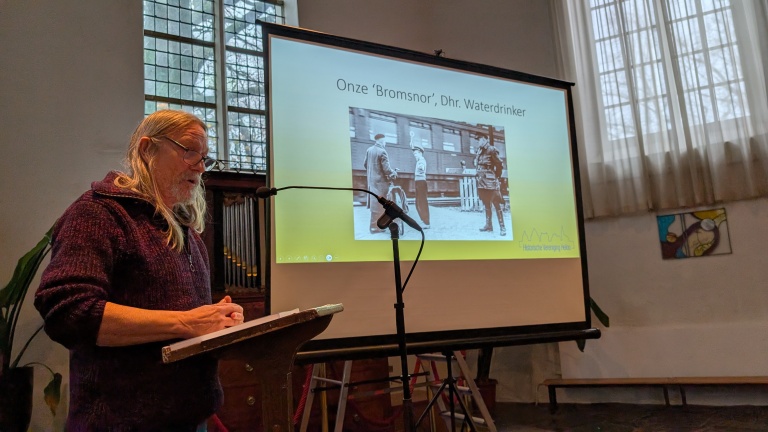Een man met lang blond haar en een baard spreekt voor een microfoon in een kerk of historische ruimte, voor een presentatie met de titel 'Onze ‘Bromsnor’, Dhr. Waterdrinker'. Op de achtergrond is een dia te zien met een oude zwart-witfoto van mensen bij een trein.