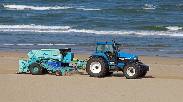 Een blauwe tractor met een strandreinigingsapparaat trekt over een zandstrand met de zee op de achtergrond.