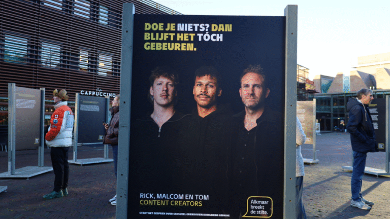 Openluchtexpositie met een groot bord met foto's van drie mannen en de tekst "Doe je niets? Dan blijft het toch gebeuren." op een plein omringd door gebouwen in Alkmaar.