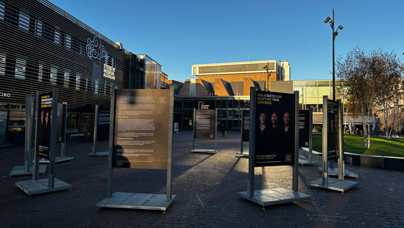 Openlucht tentoonstelling met informatieborden op een plein naast een modern gebouw met opschrift 'Studenten Mediaclowns', onder een heldere blauwe hemel.