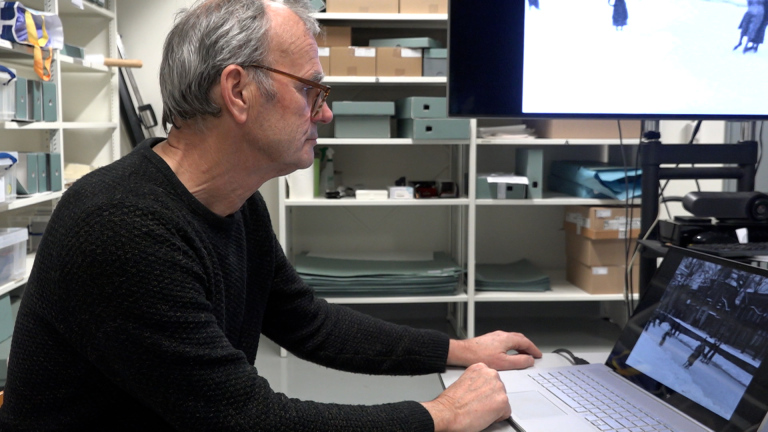 Een man met bril zit aan een bureau en kijkt naar een laptop waarop een afbeelding van een besneeuwd landschap te zien is; op de achtergrond staan planken met dozen in een archiefruimte.